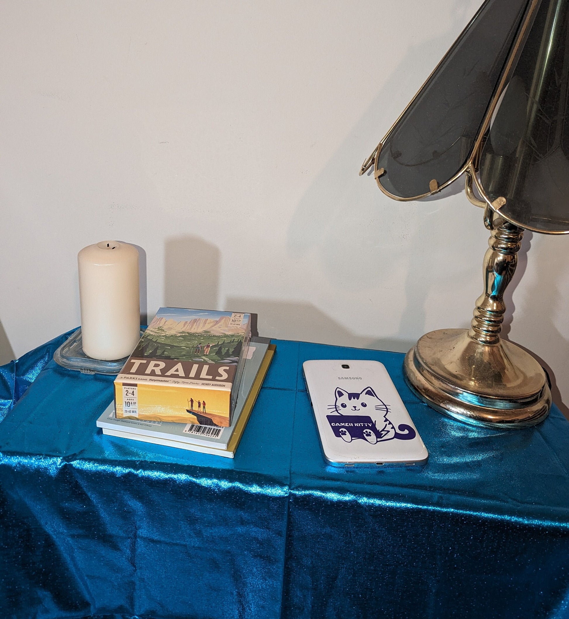 Purple vinyl cartoon kitty sitting while holding a laptop. The laptop says, &quot;Gamer Kitty.&quot; The sticker is on a Samsung tablet, along with a game, a candle, two books and a lamp on a shiny metallic teal tablecloth.